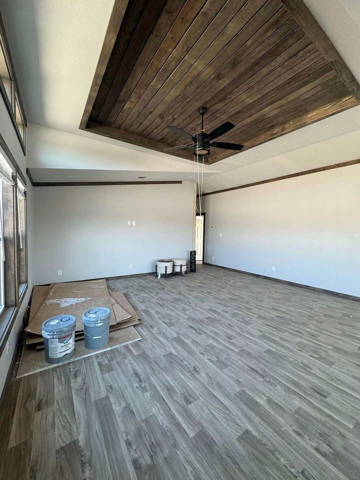 Spacious room with gray walls and wood-patterned flooring. A dark wood ceiling feature with a black fan is overhead. Paint cans and supplies on the floor suggest ongoing renovations. The atmosphere is clean and modern.