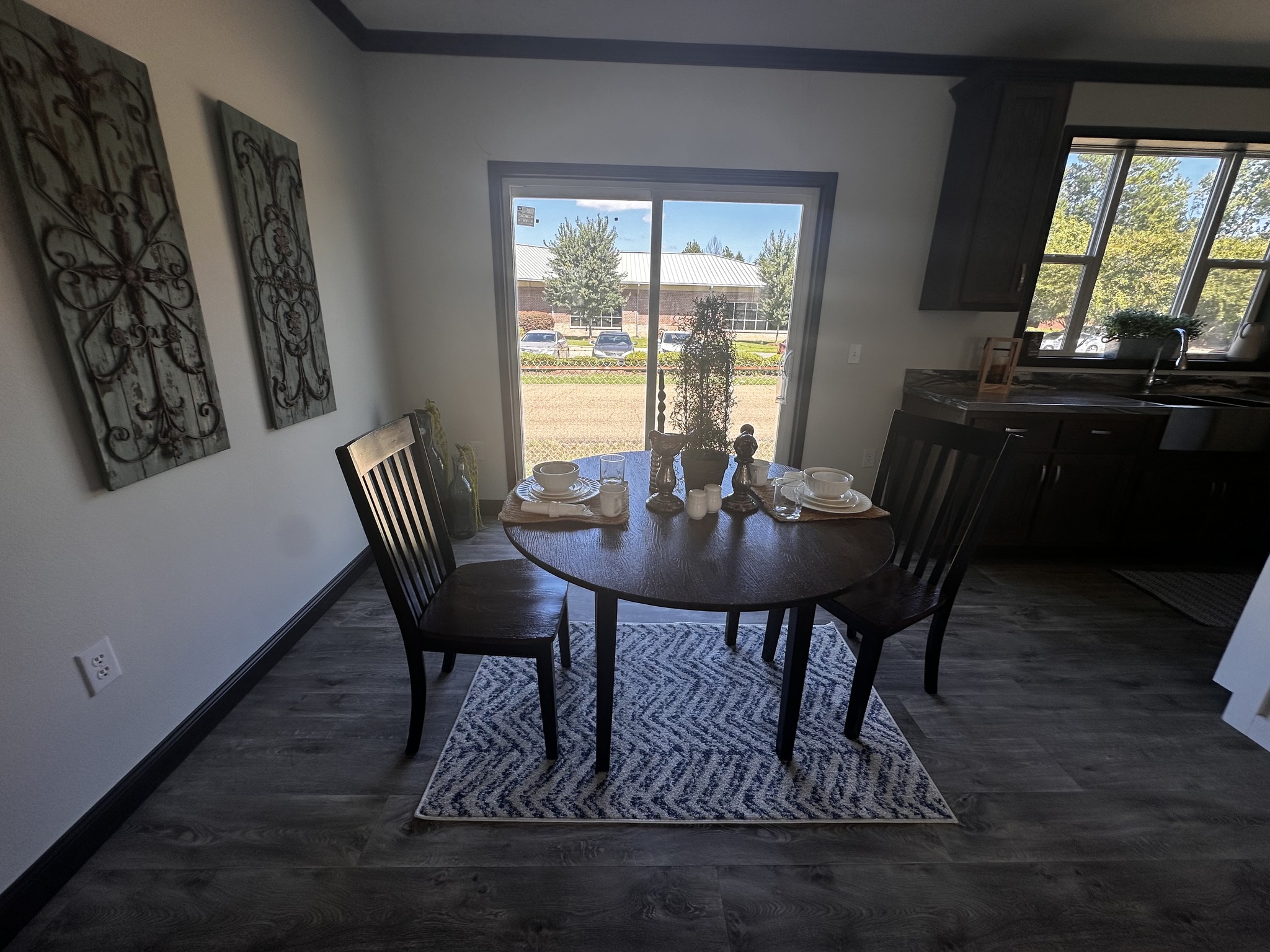 Cozy dining area with a round table set for two, on a patterned rug. Large window view of trees. Wall art and dark wooden chairs create a warm ambiance.
