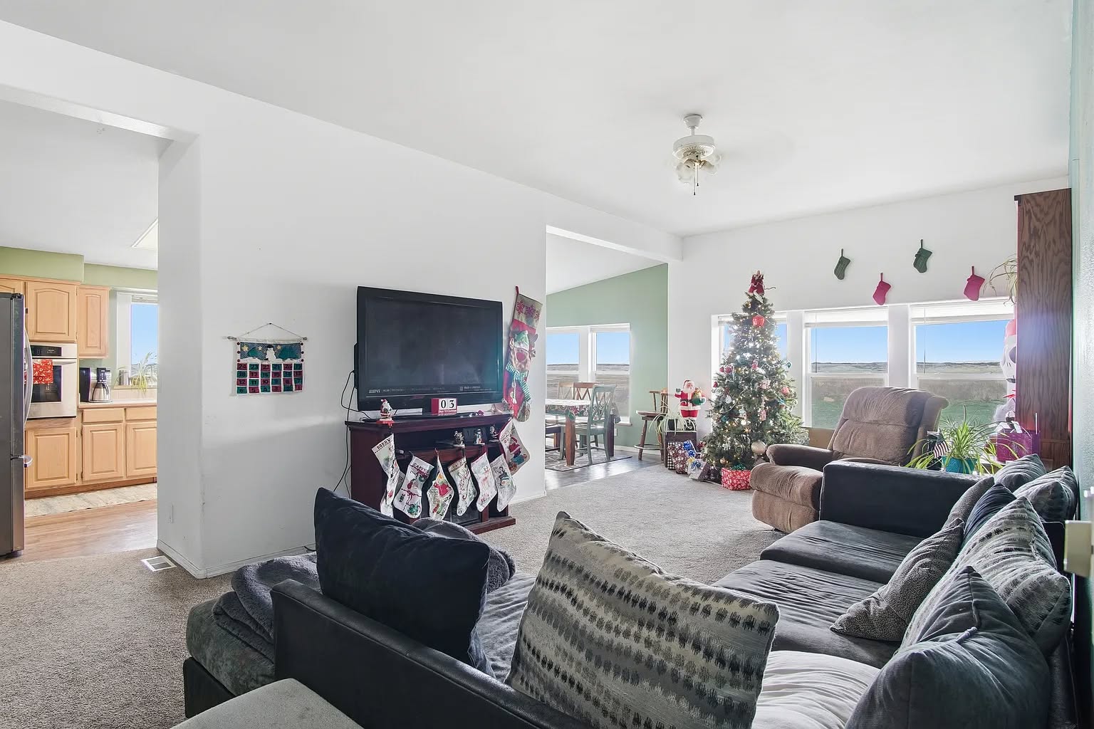 Spacious living room with a Christmas tree by a window, stockings hung on a fireplace with a TV, plush sofas, and an open view to the kitchen. Cozy holiday vibe.