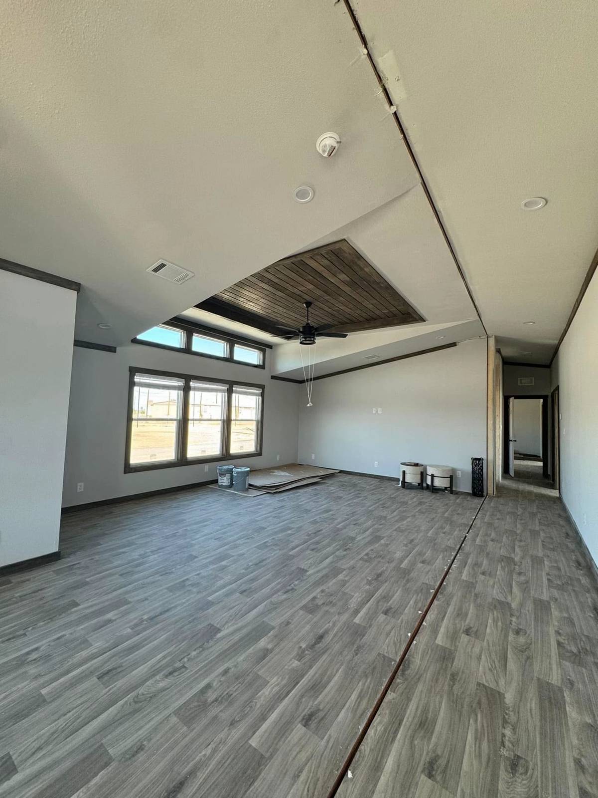 Modern, empty room with wood flooring, large windows showing a yard, and a ceiling fan. Paint cans are on the floor, suggesting renovation. Bright and spacious atmosphere.