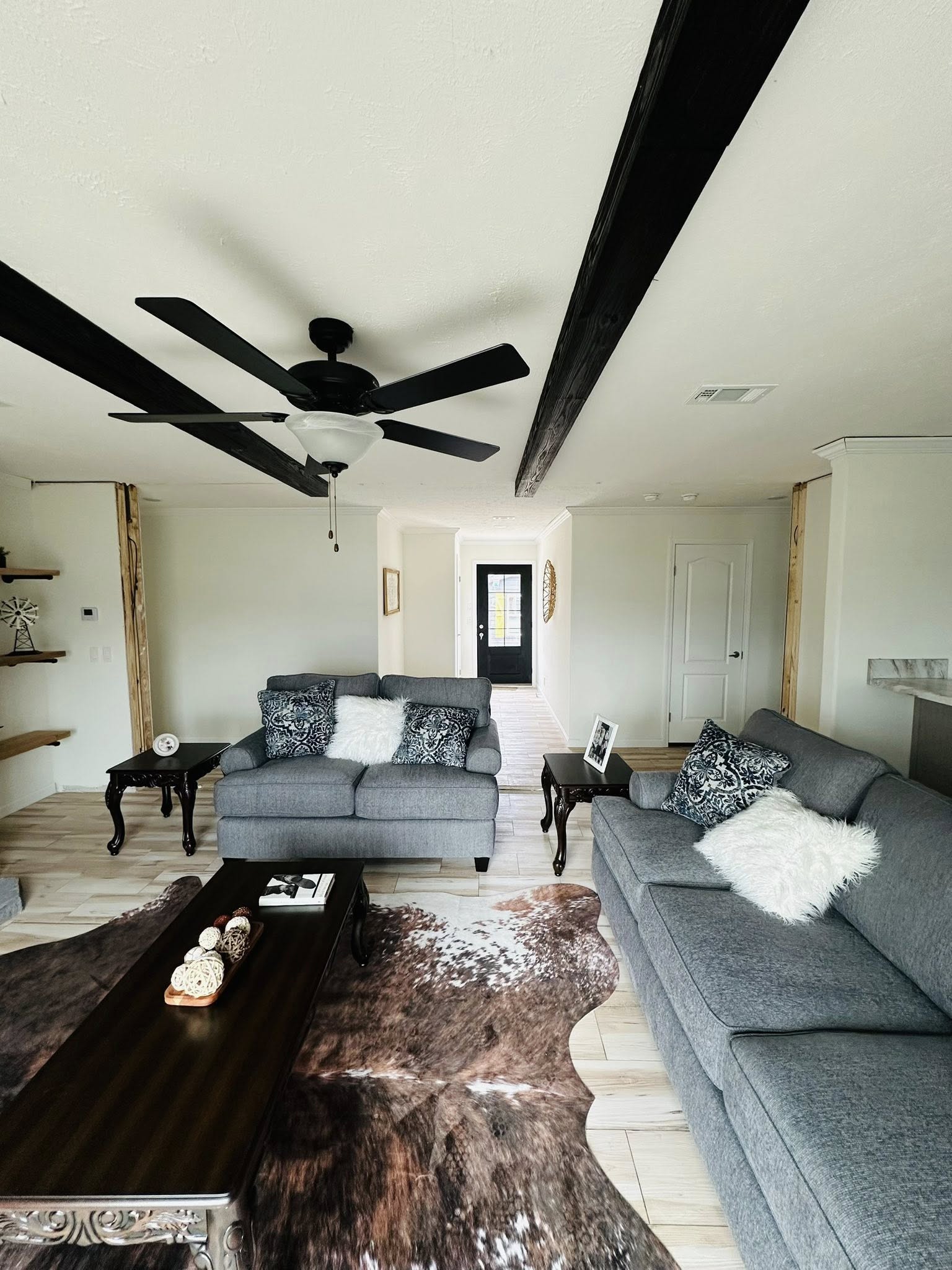 Contemporary living room with gray sofas, black ceiling fan, and wooden beams. Cozy decor includes throw pillows, textured rug, and dark coffee table.
