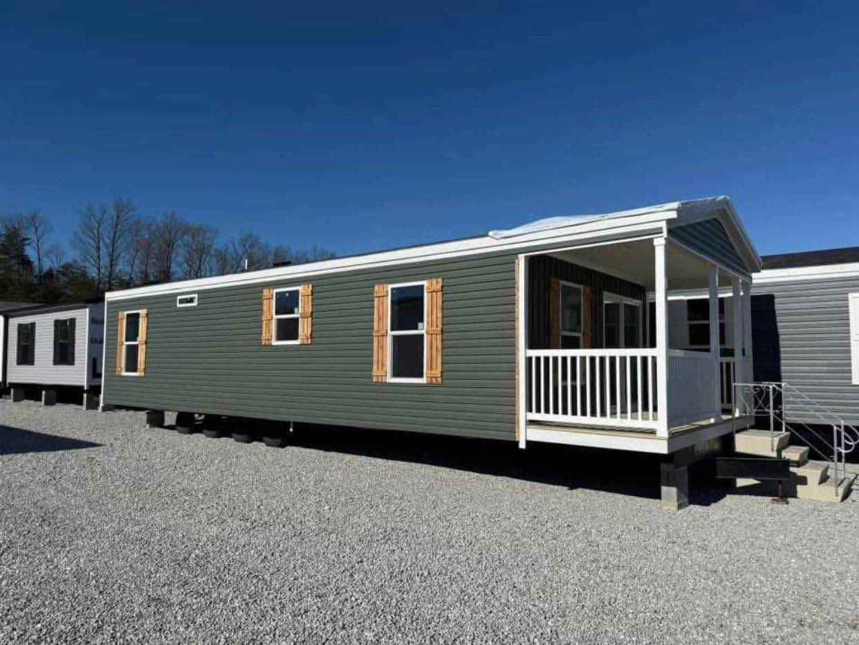Green mobile home with wooden shutters and white railing porch, set on gravel under a clear blue sky with trees in the background.
