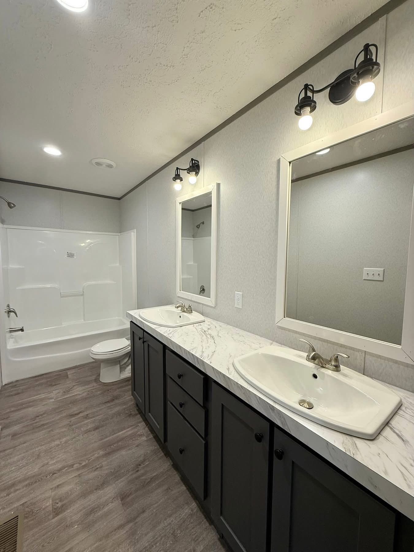 Modern bathroom with dual sinks, a white marble countertop, and dark cabinets. Two mirrors above the sinks, bathtub, and toilet in the background. Bright lighting.