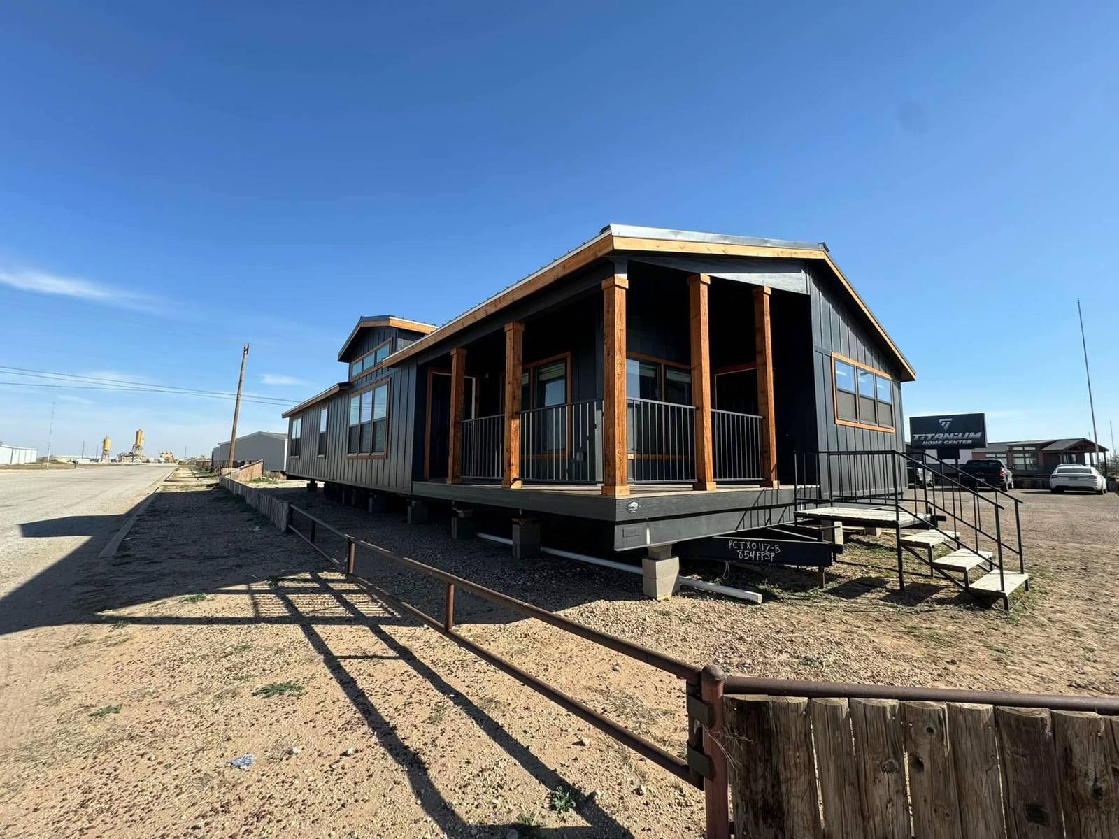 A modern, elevated modular building with wooden columns and large windows, set against a clear blue sky. It sits on a gravel and dirt lot.