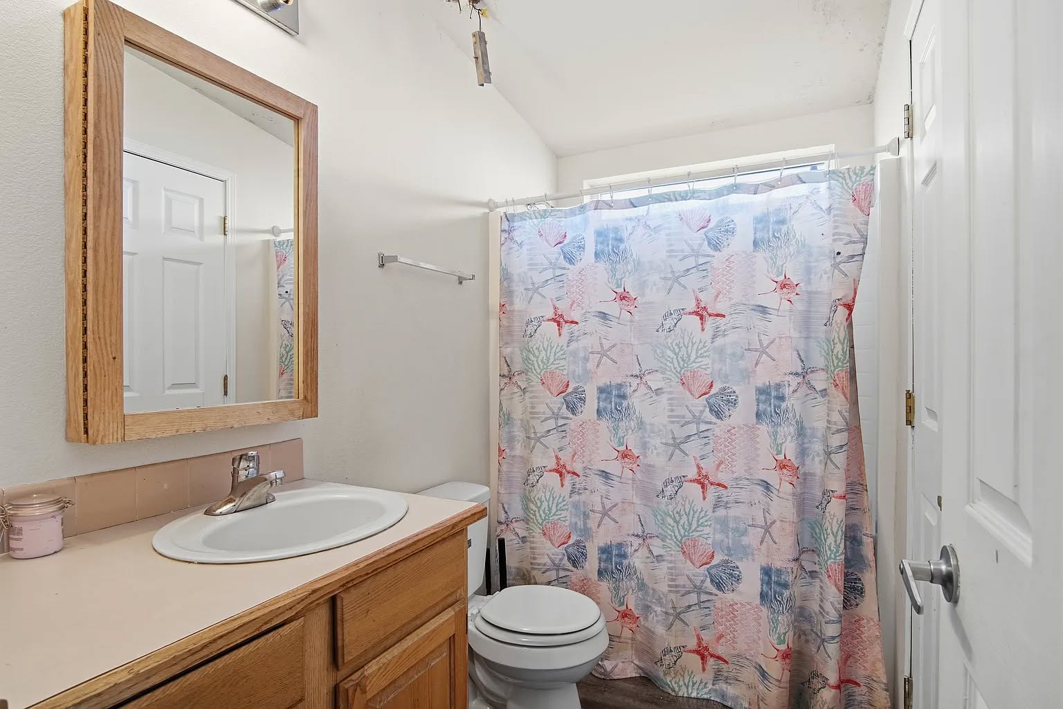 Small bathroom with a wooden vanity, mirror, and sink. A beach-themed shower curtain with starfish and seashells encloses the bathtub. Simple and tidy design.