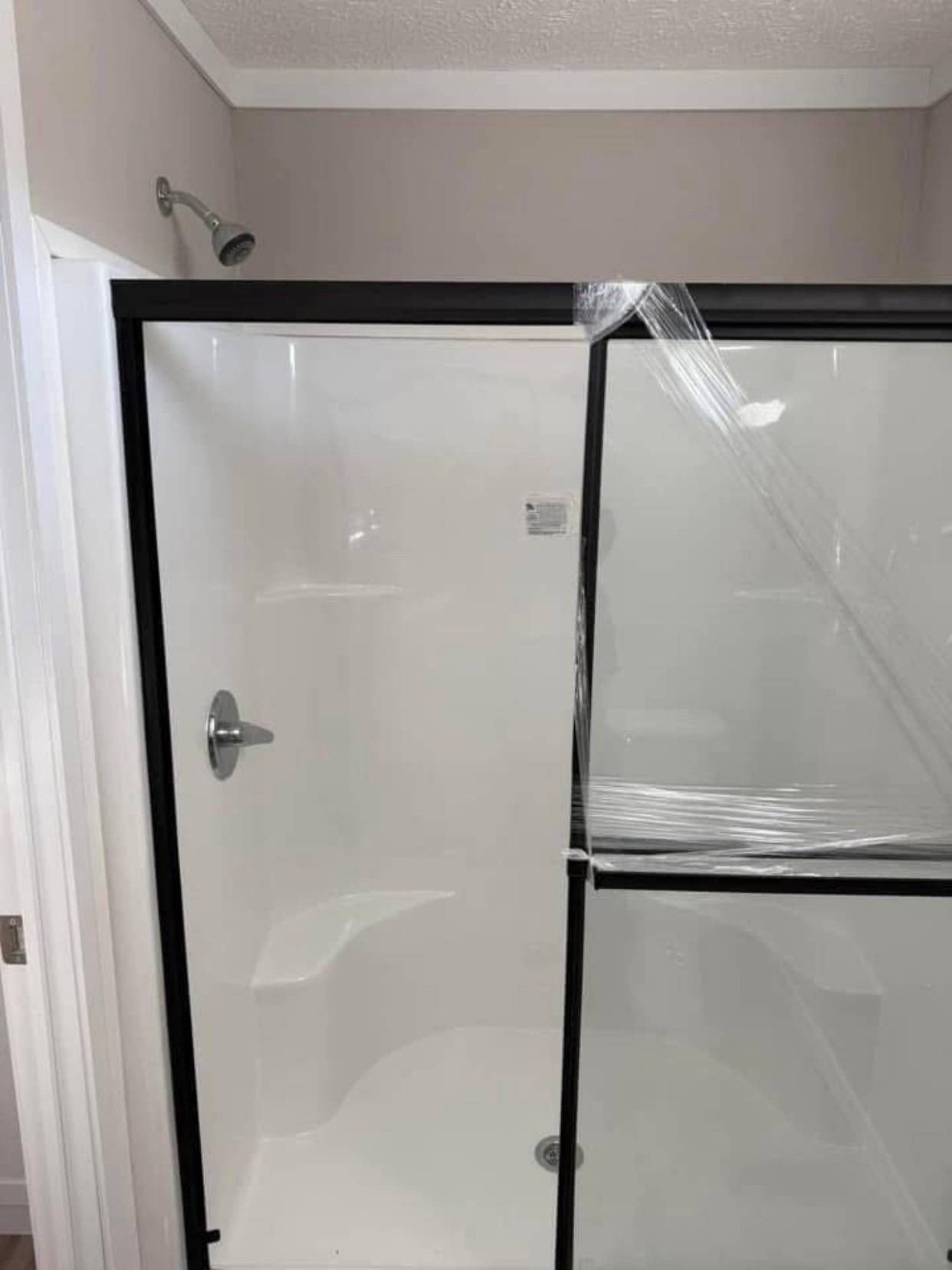 A shower enclosure with clear glass doors, half covered in protective plastic wrap. It features a silver showerhead and handle on a white backdrop.