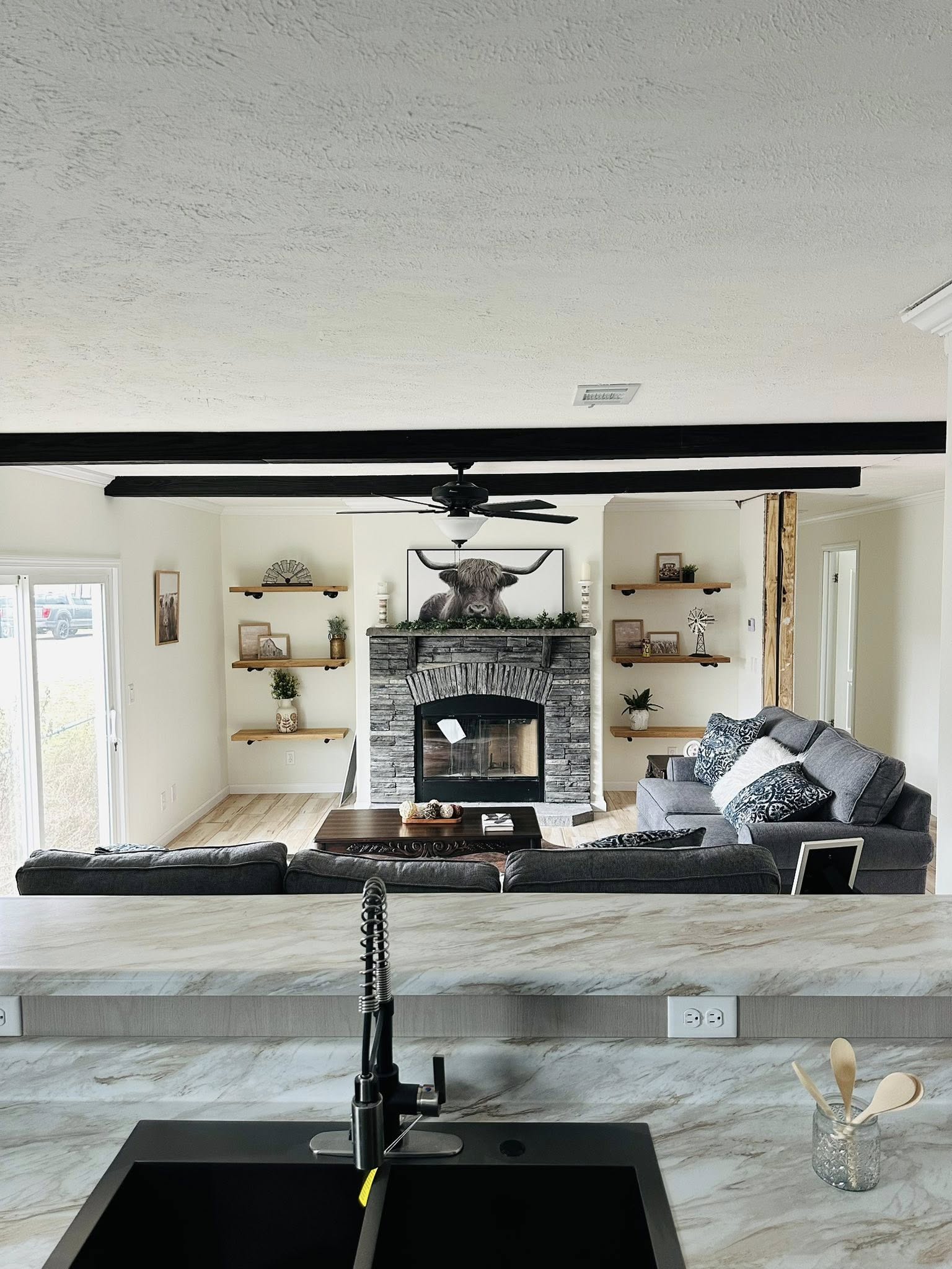Modern living room with a stone fireplace, flanked by wooden shelves with plants and decor. Gray sectional sofa faces the fireplace. View from behind a marble kitchen counter with a sink, faucet, and utensils. Bright, neutral tones create a cozy atmosphere.