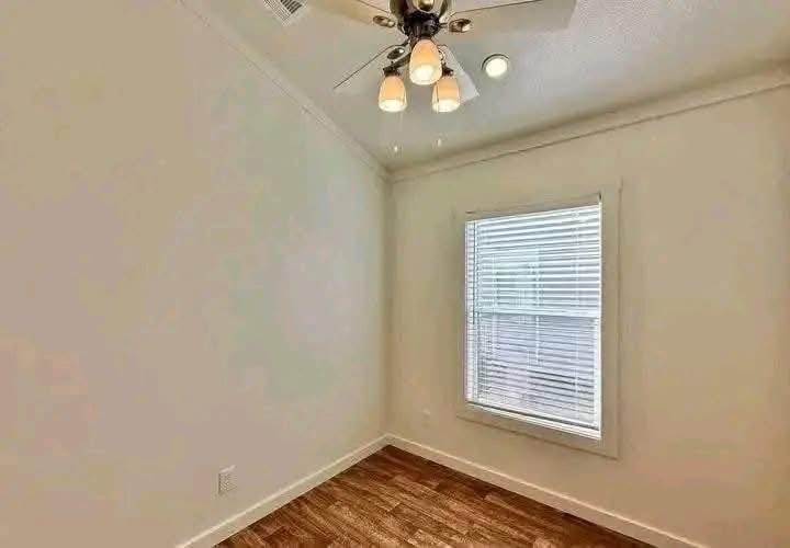 A small, empty room with light beige walls, wood flooring, a window with blinds, and a ceiling fan with lights. The atmosphere is simple and clean.