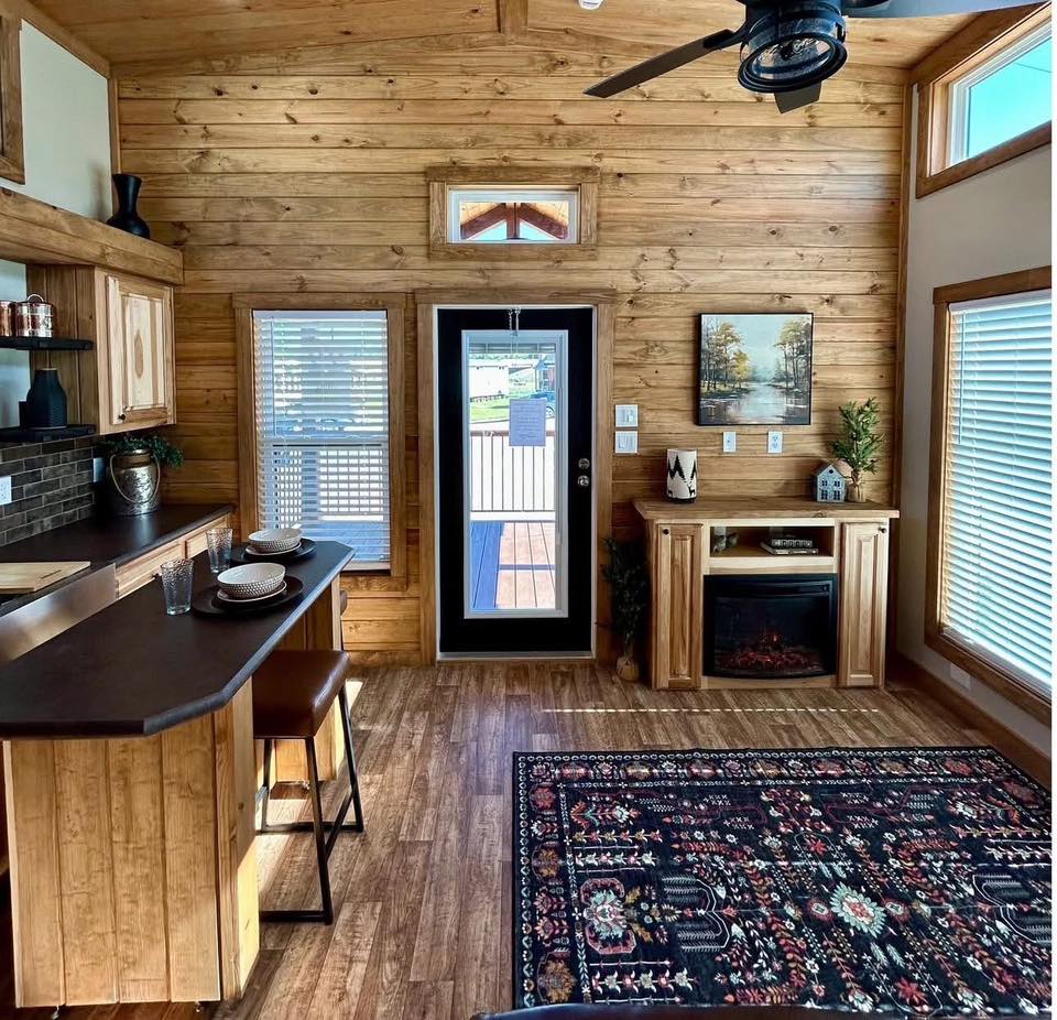 Cozy wooden interior with a high ceiling and a black door. Features a kitchen counter with stools, a patterned rug, and a fireplace. Warm, inviting atmosphere.