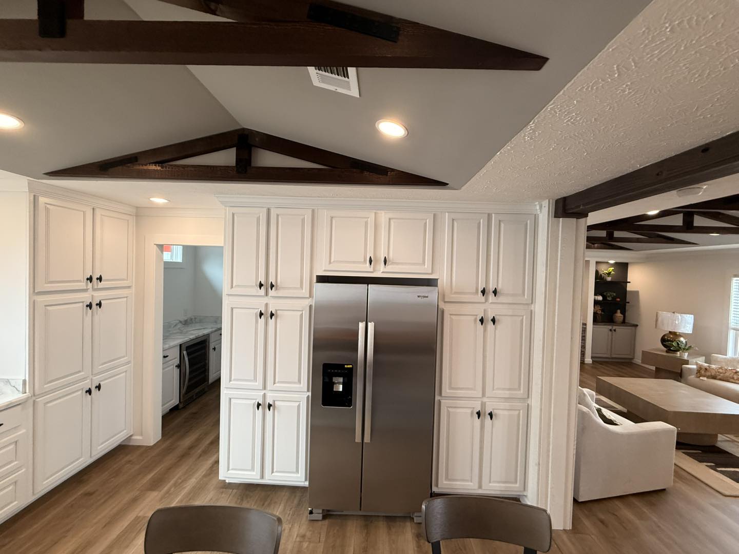 Modern kitchen with white cabinets, stainless steel fridge, wood floors, and exposed dark wood ceiling beams. Open to a cozy living space with a warm ambiance.
