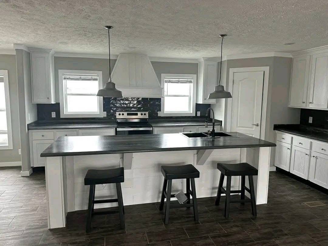 Modern kitchen with a monochrome tone, featuring stainless steel appliances, dark countertops, white cabinets, and three black stools by the island.