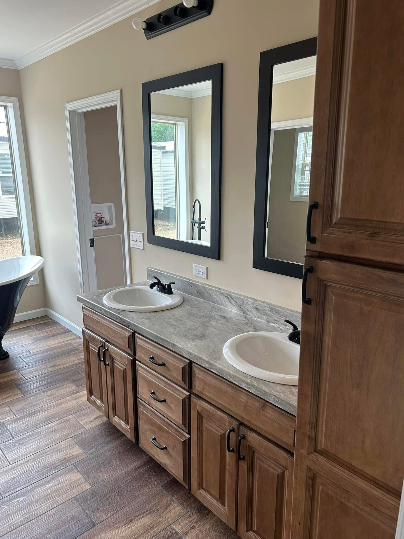 Bathroom interior with a double-sink vanity featuring marble countertops and dark faucets. Two rectangular mirrors hang above. Natural light from a window softly illuminates the space, which features light beige walls and wood flooring for a cozy ambiance.