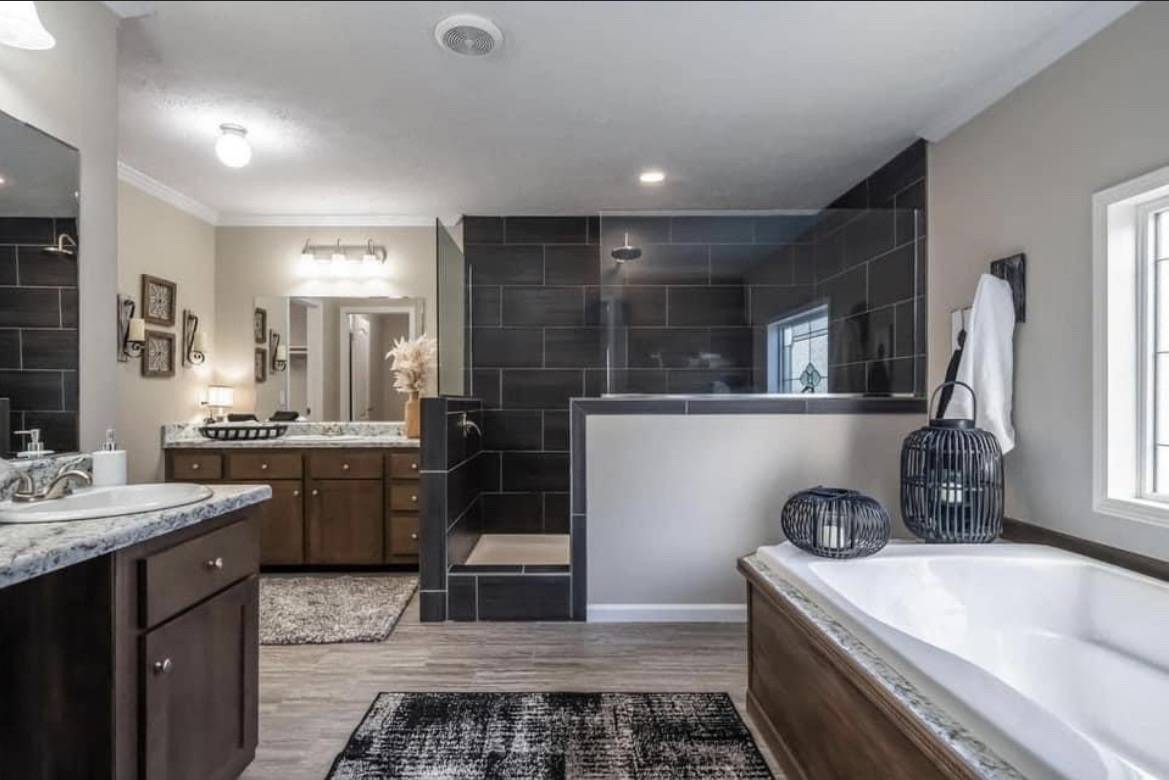 Spacious bathroom with dark tile walk-in shower, a white bathtub with wood accents, dual sinks on granite countertops, and decorative lanterns.