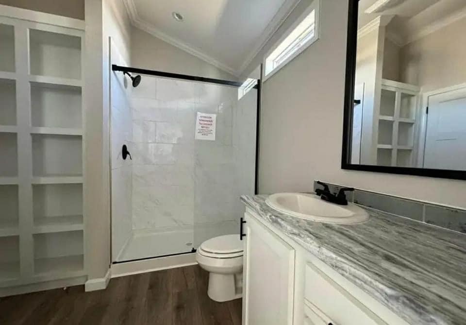 Modern bathroom with a glass-enclosed shower, wooden flooring, and a marble countertop. Shelving unit and subdued lighting create a clean, minimalist feel.