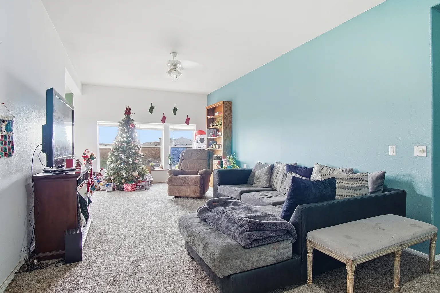 A cozy living room with a teal accent wall, featuring a large sectional sofa and a plush recliner. A decorated Christmas tree and stockings add festive cheer near the window, with wrapped gifts beneath. A flat-screen TV sits on a stand, and natural light brightens the room through large windows.