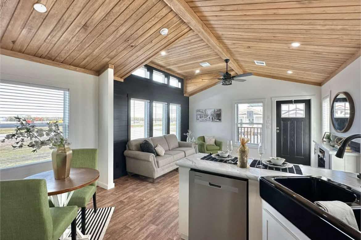 Modern living room with wood-paneled vaulted ceiling, large windows, gray sofa, green chairs, and an open kitchen with island. Bright and inviting space.