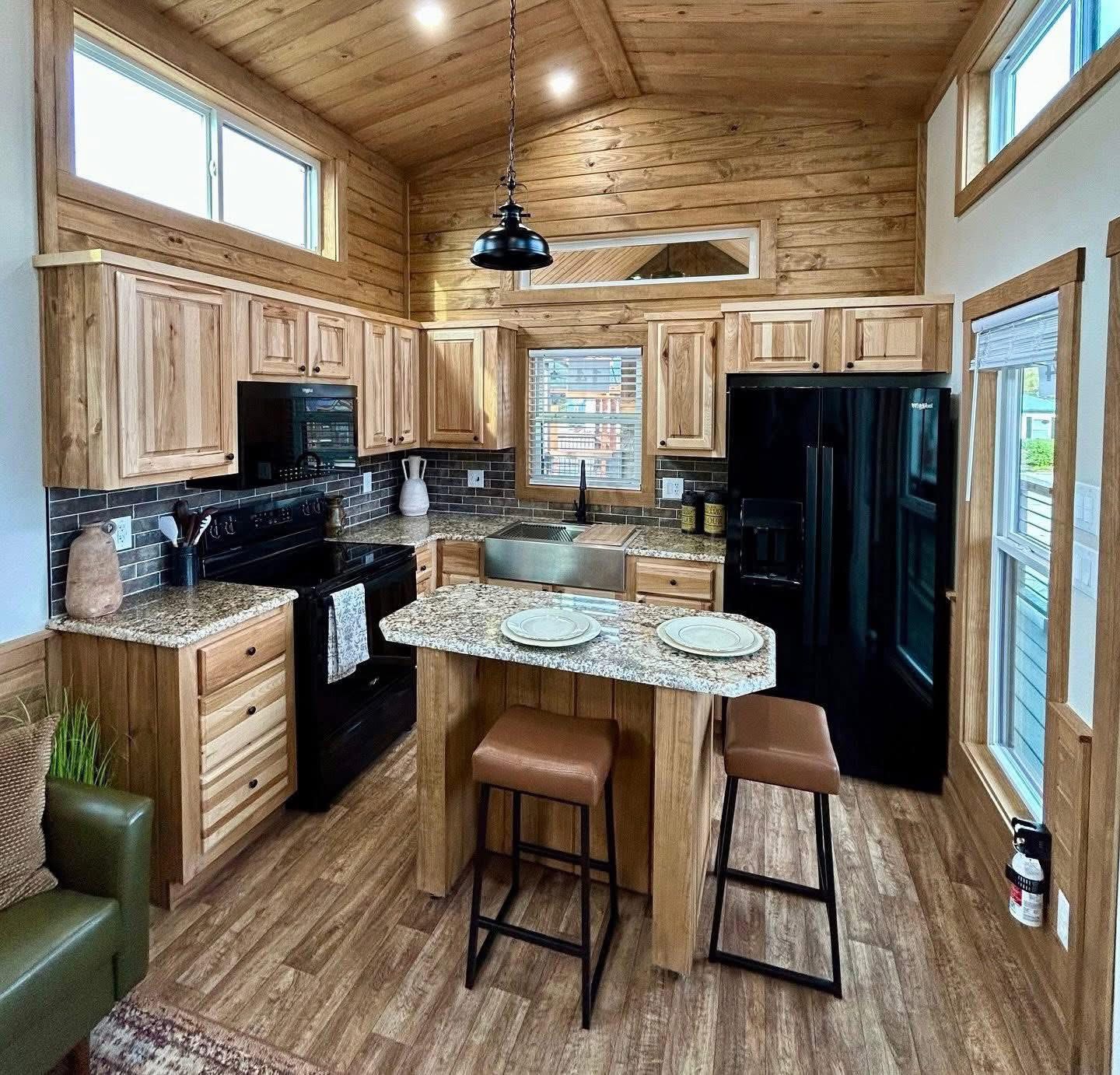 Cozy kitchen with wooden cabinetry and ceiling, granite countertops, and an island with two stools. Large windows and black appliances add contrast.