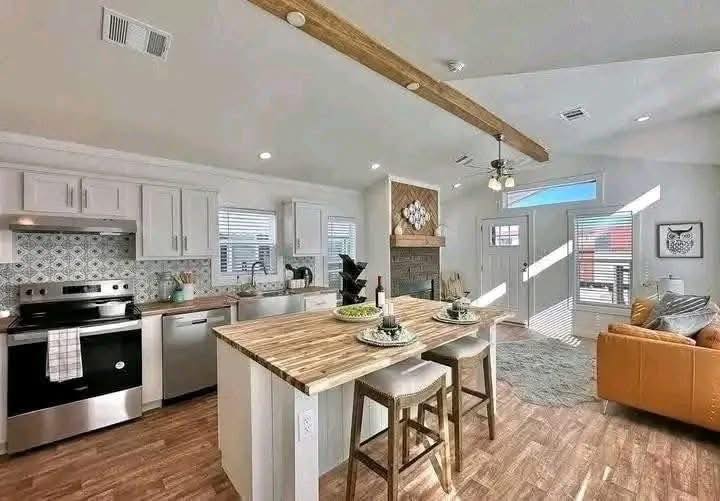 Bright, modern kitchen and living area with wood flooring, a central island with stools, white cabinets, stainless steel appliances, and a cozy seating space.
