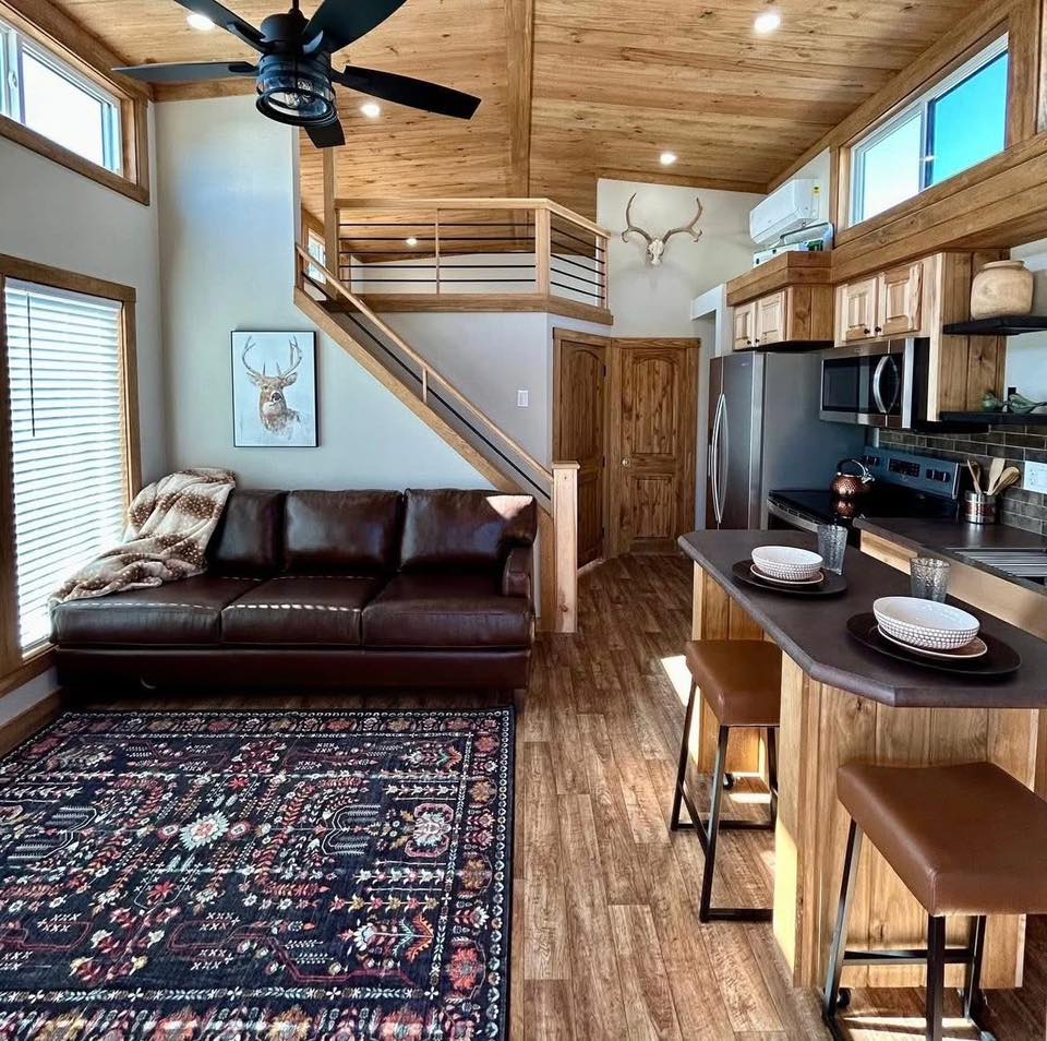 Cozy living room with a brown leather sofa, patterned rug, and deer artwork. Wooden loft and kitchen, featuring modern appliances and barstools. Warm ambiance.