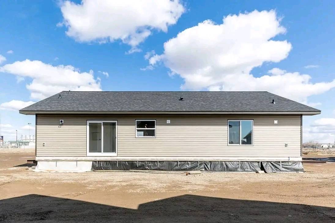 Simple beige house with a dark roof and plastic-covered foundation sits on a dirt lot. Bright blue sky with scattered clouds in the background.