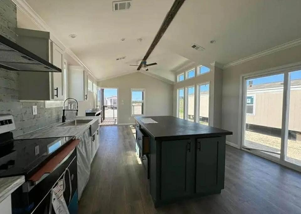Bright kitchen with modern design, featuring gray cabinets, a dark island, stainless steel sink, large windows, and wooden floor, creating an airy feel.