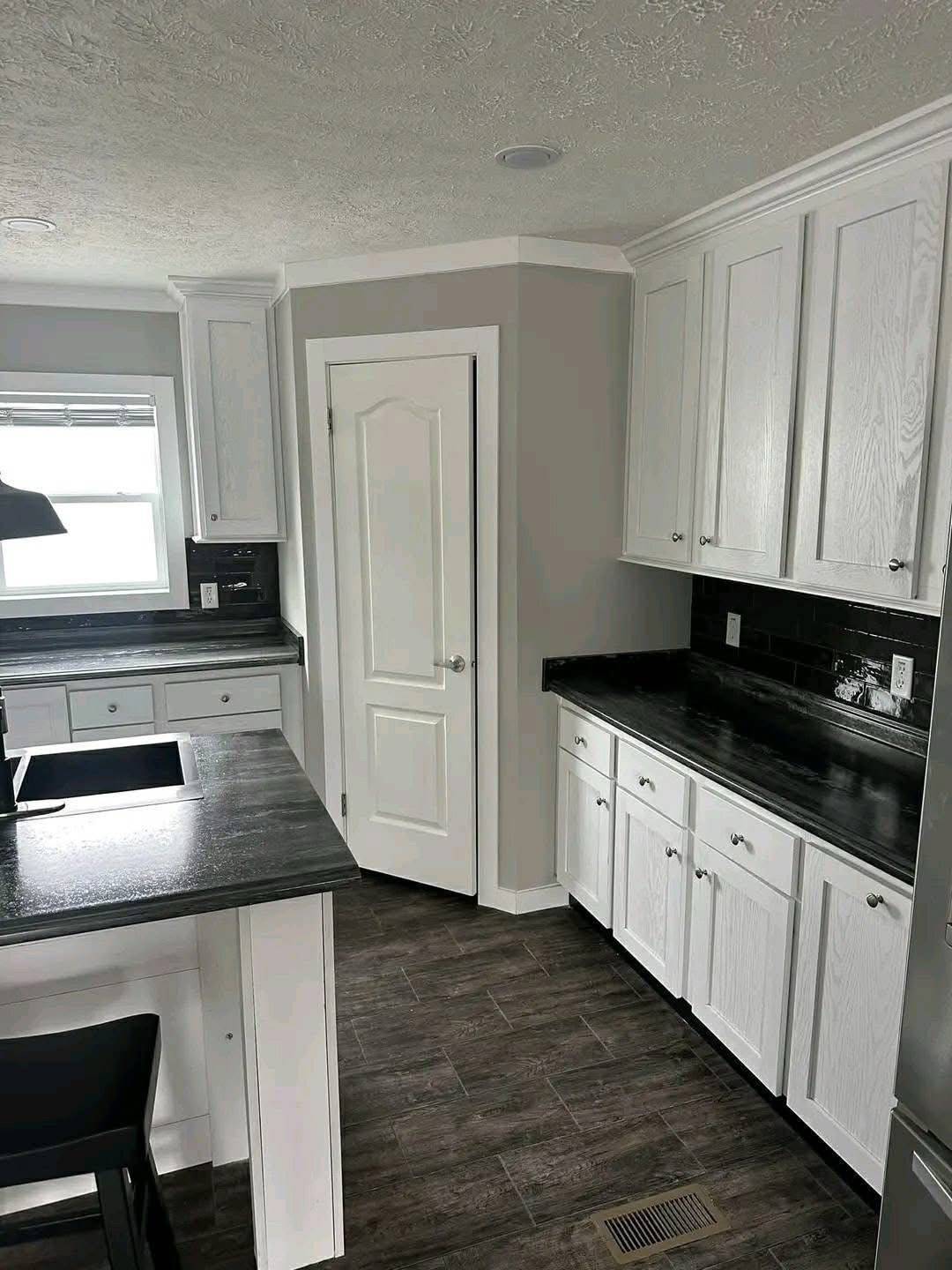 Modern kitchen with white cabinets, dark countertops, and a gray tiled floor. A central island has a sink; window brings in natural light. Calm, sleek atmosphere.