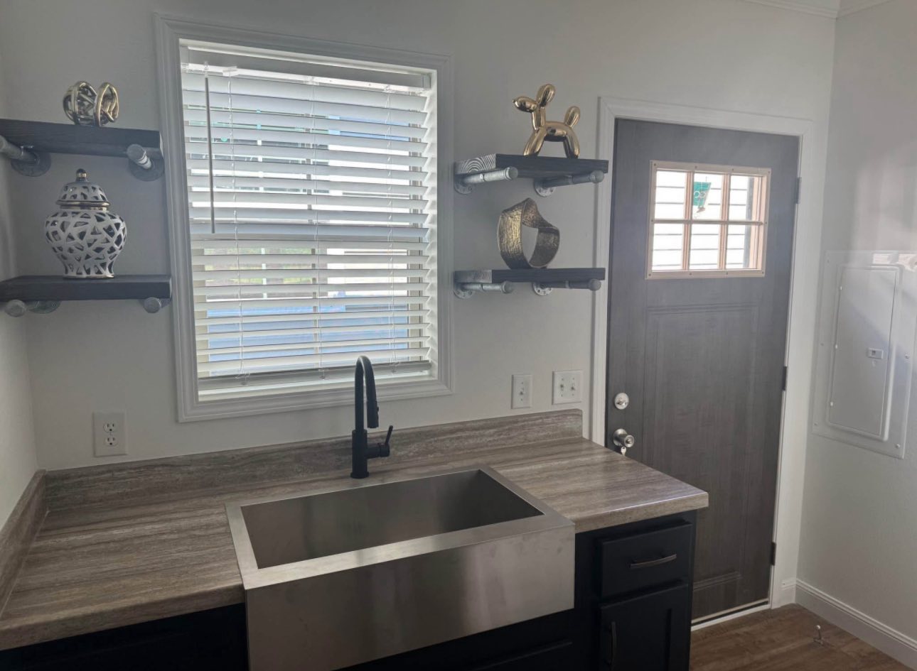 Compact kitchen space with a stainless steel sink, black faucet, and wooden countertops. Wooden shelves hold decorative items. Light streams through a window with blinds, creating a calm atmosphere. Door on the right with a grid window.