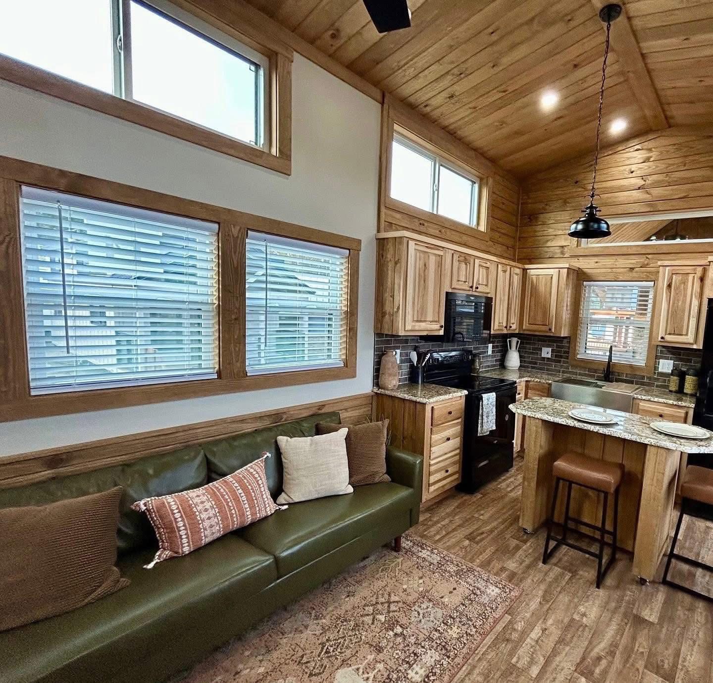 Cozy cabin interior with wooden walls and ceiling, green sofa with pillows, and a kitchen area featuring wood cabinets, granite island, and pendant light.