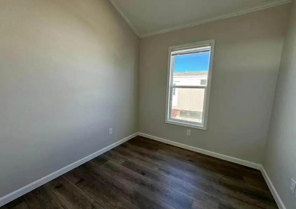 A small empty room with light beige walls and dark wood floors. A single window lets in bright natural light, showing a blue sky and building outside.