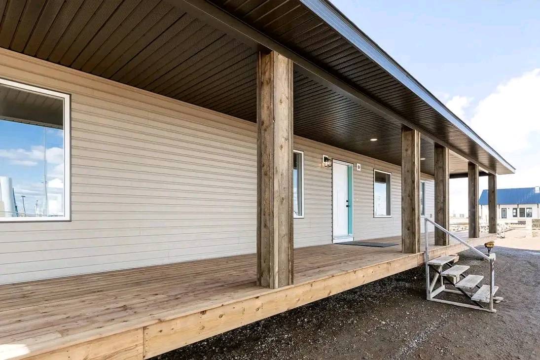 A wooden porch with large beams supports a house with light siding and a teal door. Steps lead to the entrance, conveying a rustic and welcoming feel.