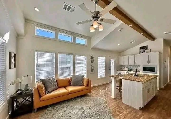 Modern living room and kitchen with high ceilings. Features a tan sofa, white cabinetry, wooden island, ceiling fan, and large windows. Bright, airy atmosphere.