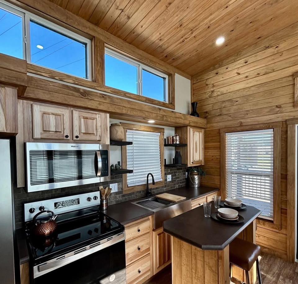 Cozy wooden kitchen with a high ceiling, sleek appliances, and a breakfast bar set for two. Warm lighting and natural tones create a rustic, inviting feel.
