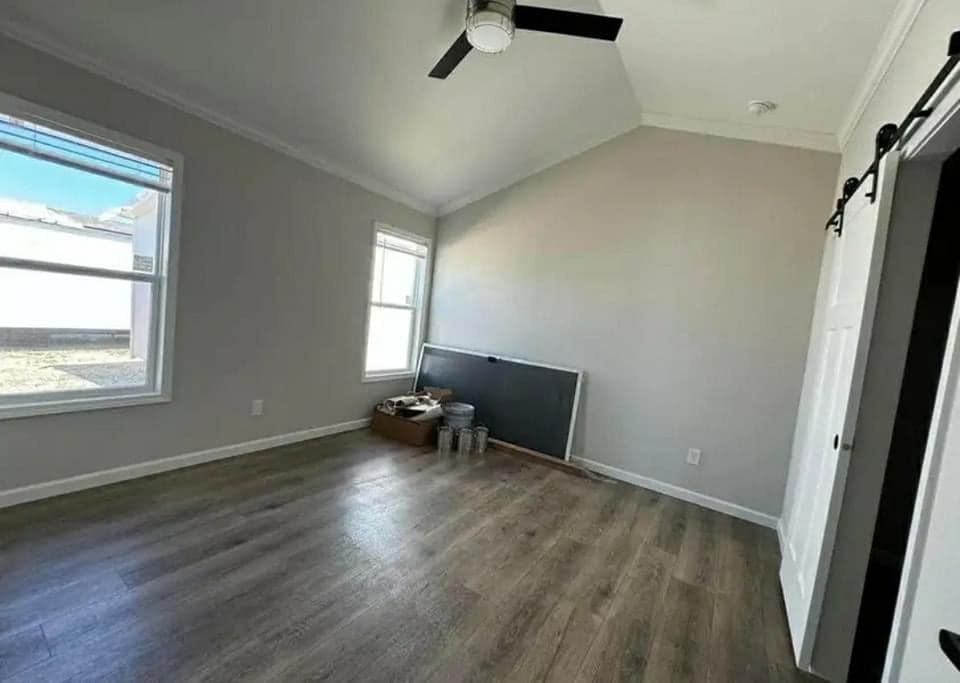 A spacious, empty room with light gray walls and wooden floors. Two large windows let in natural light. Painting supplies are by a wall, giving a calm, unfinished feel.