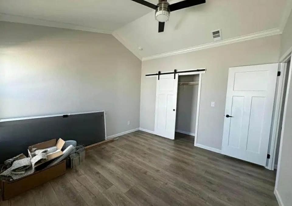 Minimalist room with light wooden floors, a ceiling fan, and gray walls. A closet with sliding barn doors is on the right. A cardboard box and materials lie in the corner, adding an unfinished feel.
