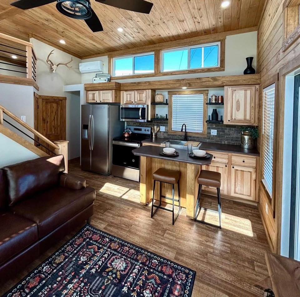 Cozy cabin living room with wooden accents. Includes a modern kitchen, brown leather sofa, patterned rug, and antler decor, creating a warm atmosphere.