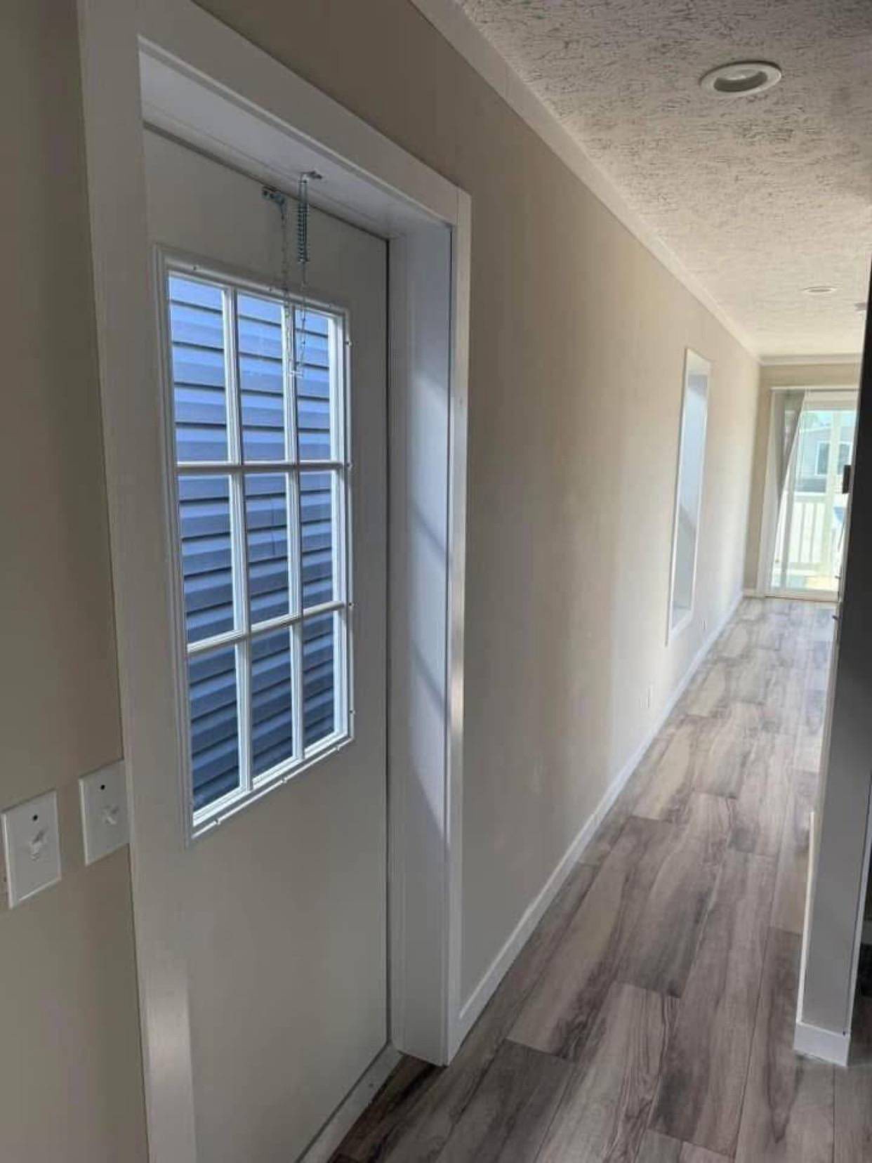 Narrow beige hallway with wood floor, featuring a windowed door to the left revealing blue siding, leading to bright glass doors at the end.