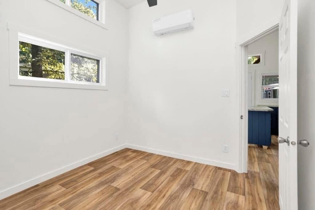 A bright, empty room with white walls, light brown wood flooring, and two windows showing green trees outside. An air conditioner is mounted high on the wall, and a door opens to a room with a blue cabinet, conveying a minimalist, fresh atmosphere.