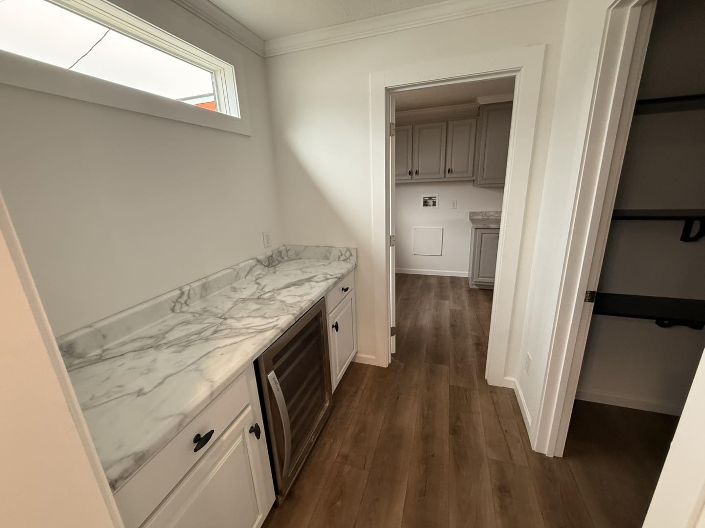A narrow laundry room with a marble countertop, wooden flooring, and white cabinets. A small wine fridge is beneath the counter. There's an open door leading to another room with cabinetry. The setting conveys neatness and modern elegance.