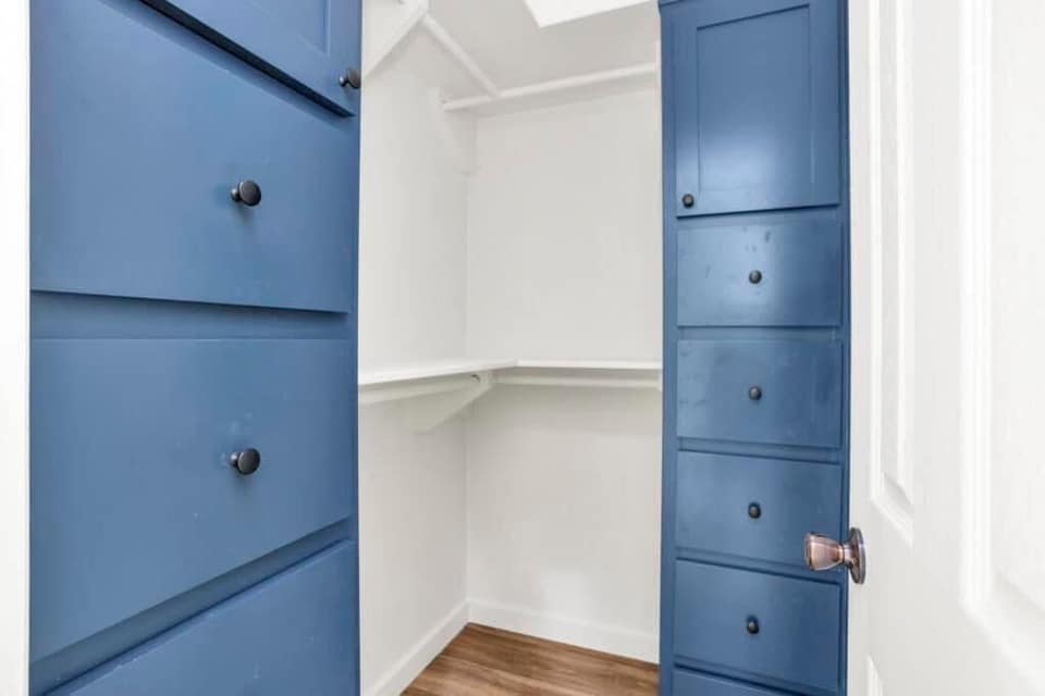 Small, white walk-in closet with blue cabinets and dark knobs on the left, and hardwood flooring. Clean, organized, and modern aesthetic.