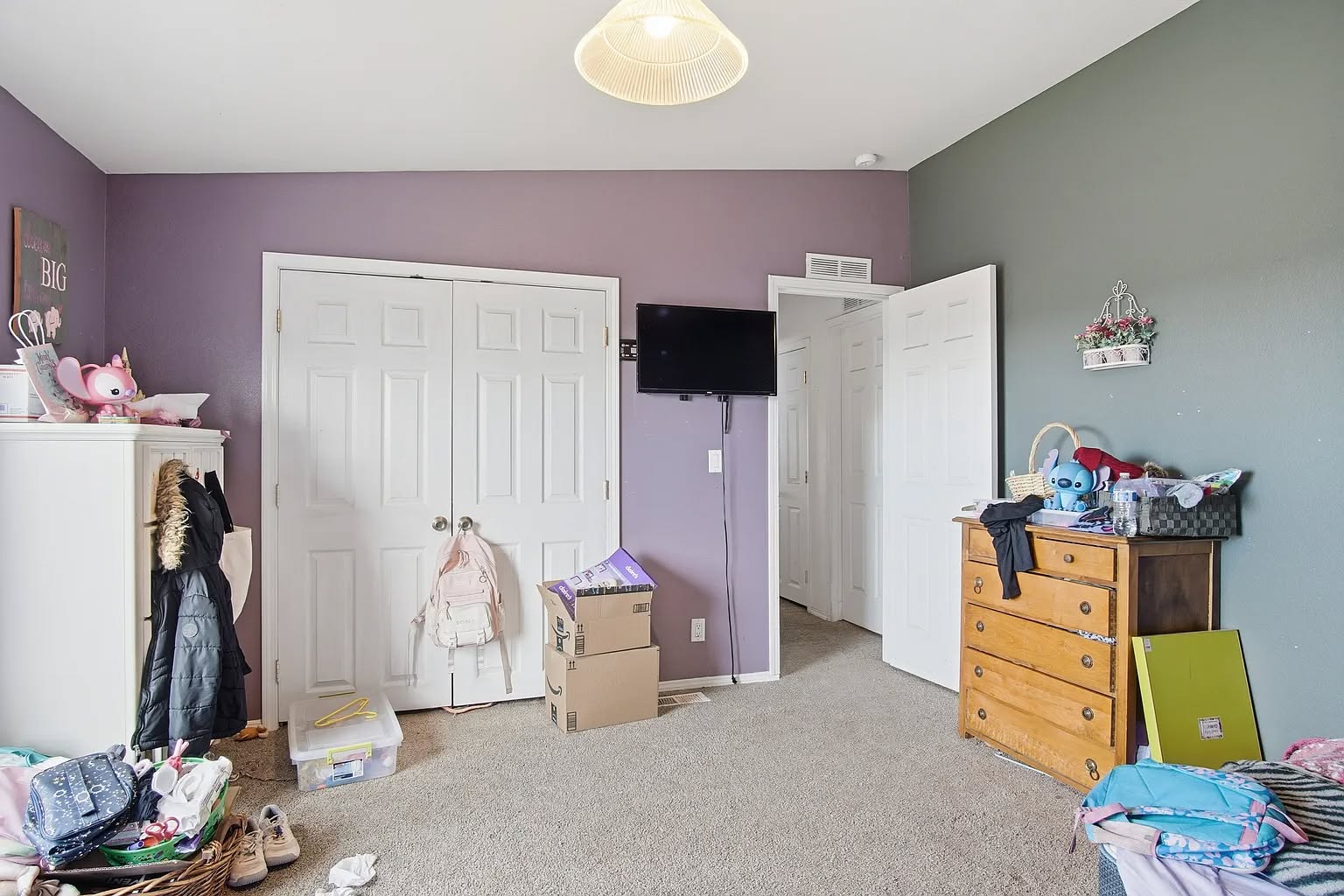 A child's room with purple and green walls, cluttered with clothes, toys, boxes, and a dresser. Cozy but messy atmosphere with a wall-mounted TV.