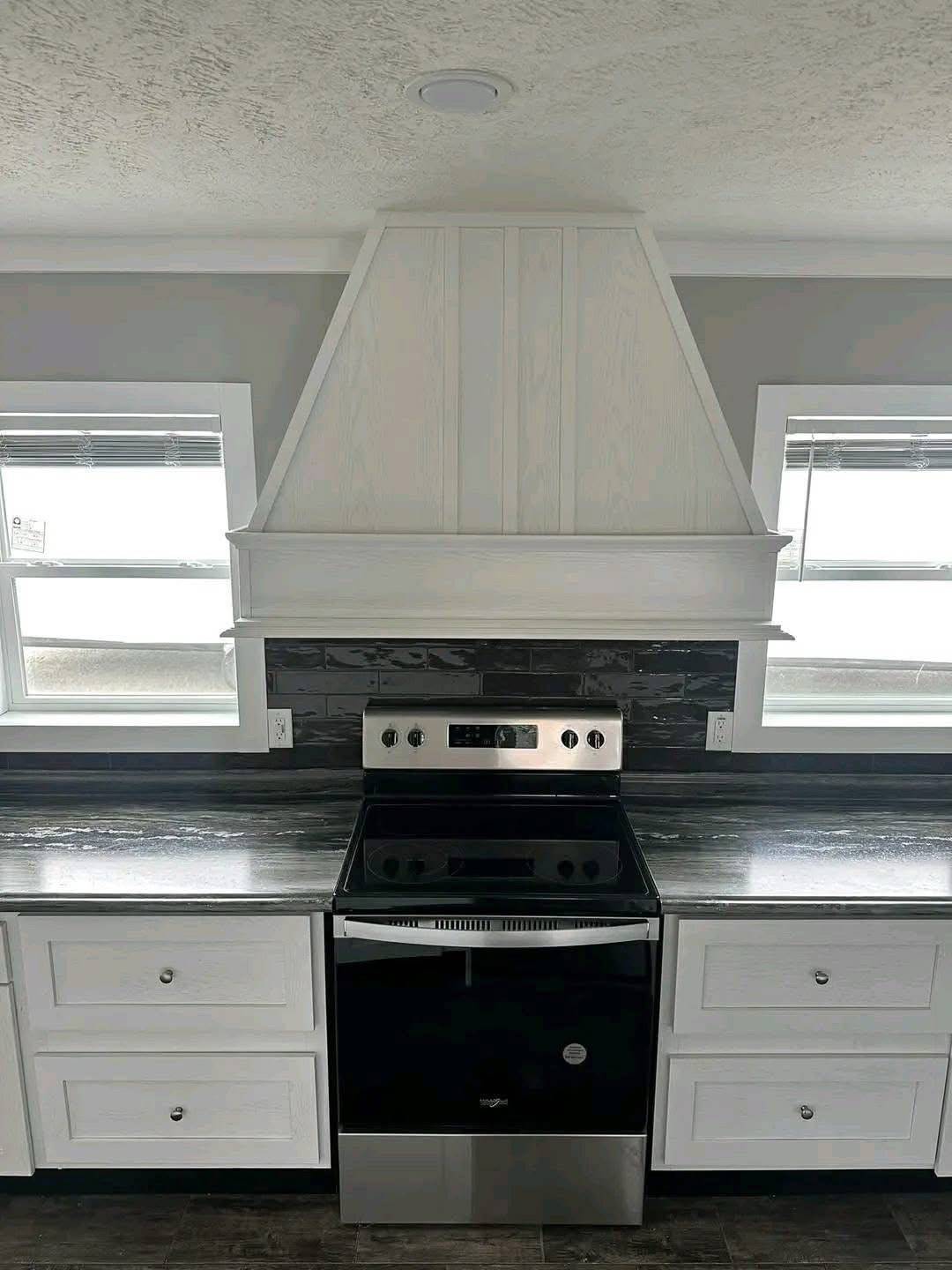 Modern kitchen with sleek black stove, white cabinets, and a large white range hood. Windows on both sides create a bright, clean ambiance.