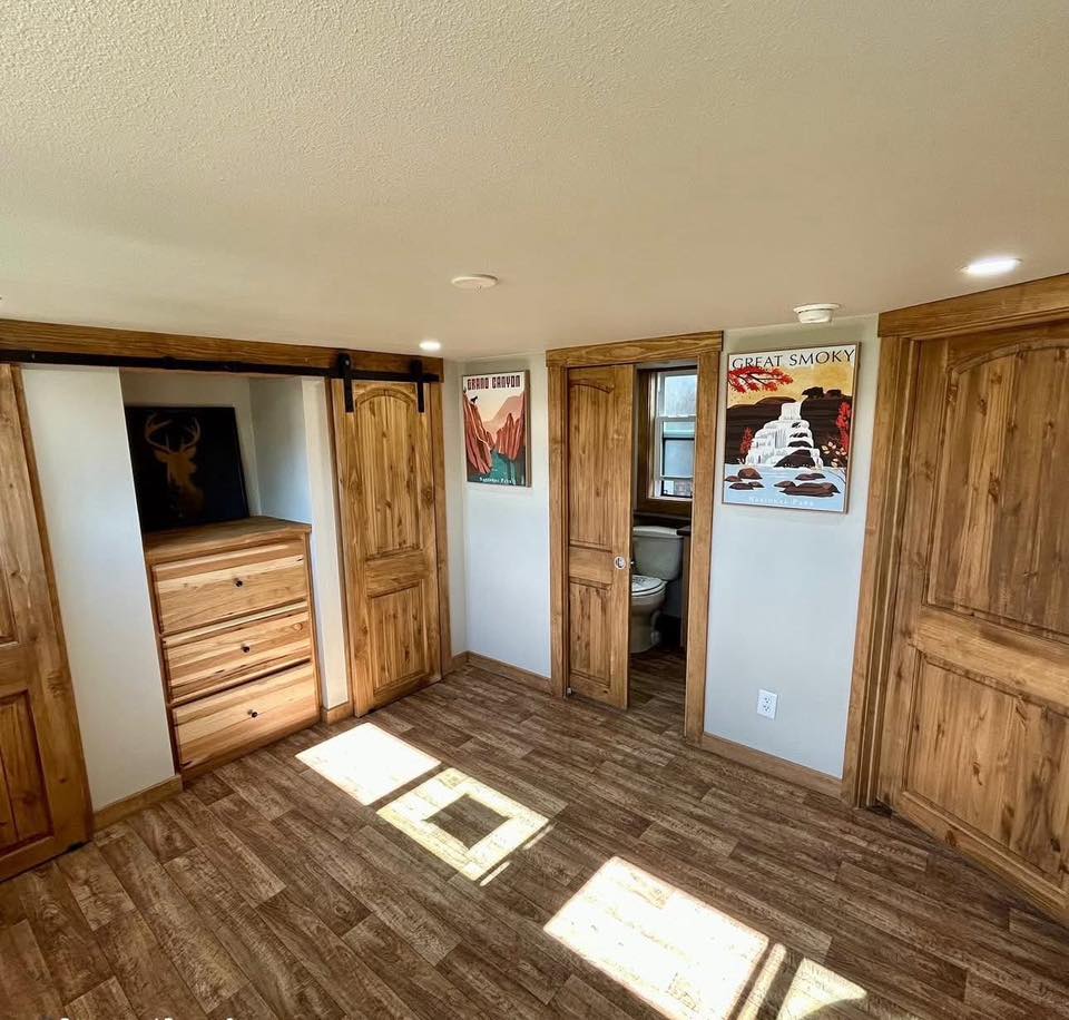 A cozy room with wooden floors and light wood doors. Two posters hang on white walls, and sunlight streams through a window, casting patterns on the floor.