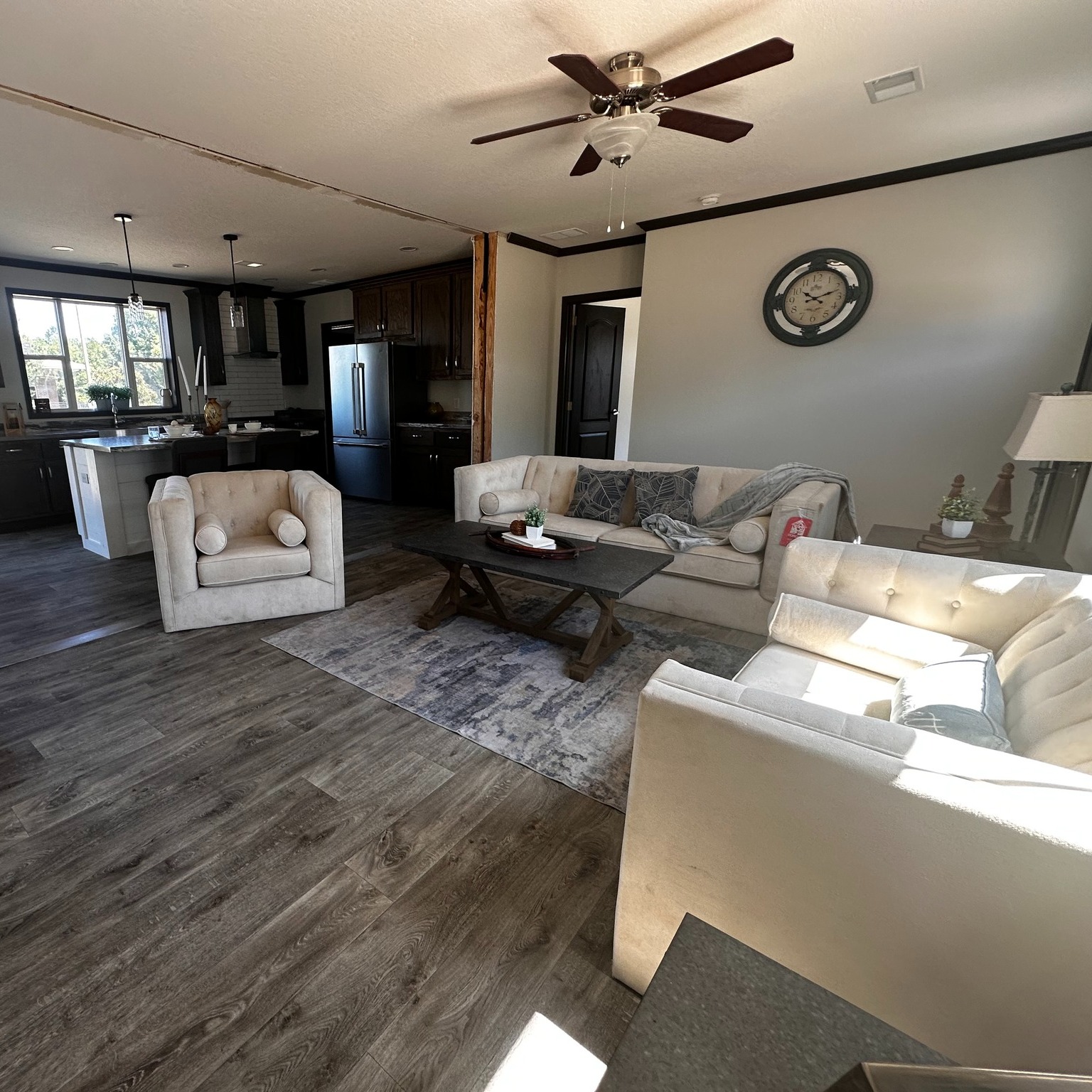 Bright living room with cream sofas, a wooden coffee table, and a ceiling fan. Open kitchen with dark cabinets is visible. Cozy, inviting atmosphere.
