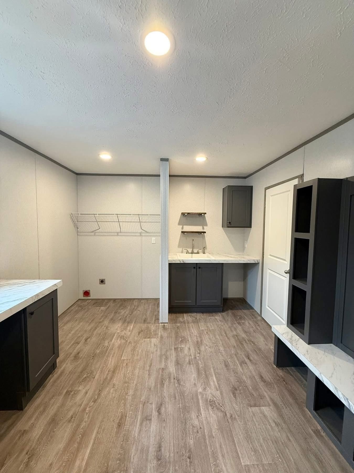 Laundry room with wood flooring, overhead lighting, and light gray walls. Features dark cabinets, a countertop sink, open shelving, and a white door.