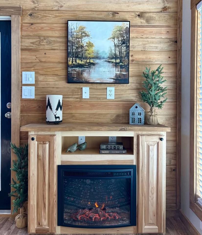 A cozy living room with a wooden accent wall features an electric fireplace, a nature painting above, and decor items like a small tree and lantern, evoking warmth.