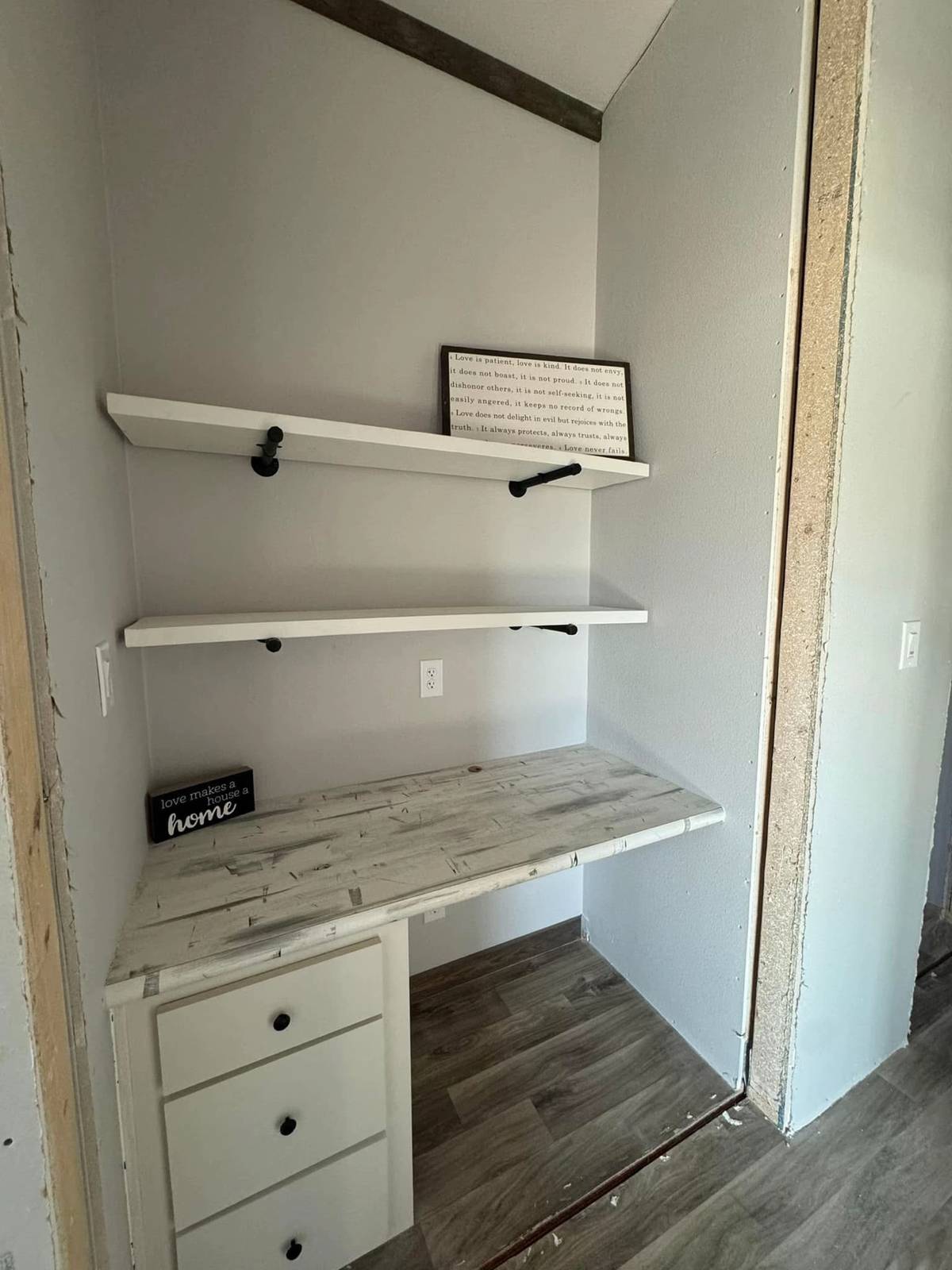 Cozy nook with weathered white desk, three drawers, and two wooden shelves. A sign reads "love makes a house a home," creating a warm atmosphere.