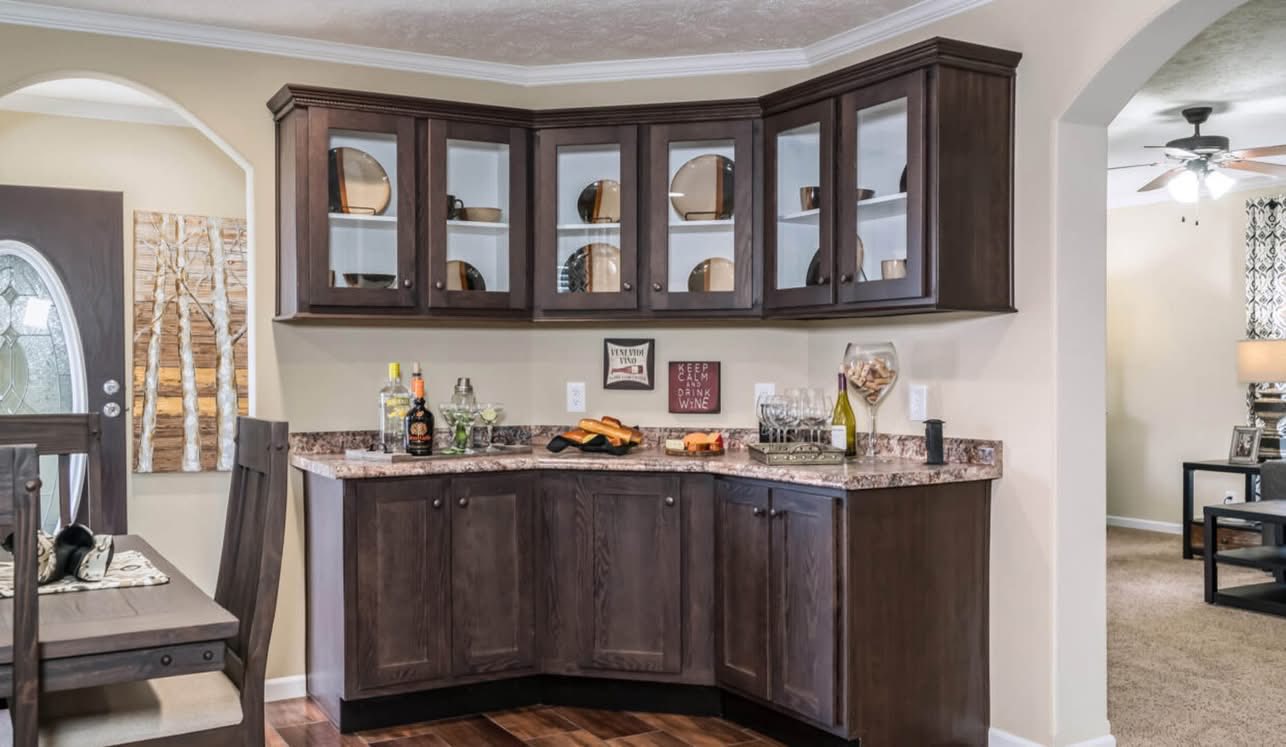 A cozy home corner bar features dark wood cabinets topped with glasses, bottles, and decor. The room feels warm and inviting, showcasing a homey atmosphere.