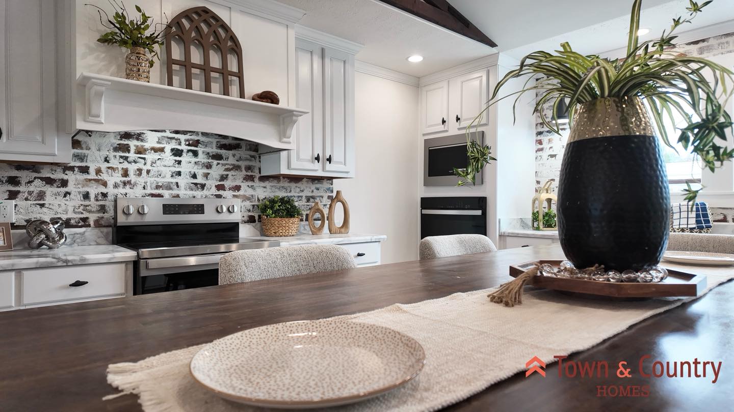 Warm kitchen with rustic charm features white cabinets, brick backsplash, and greenery. A wooden table centerpiece adds a cozy, inviting feel.