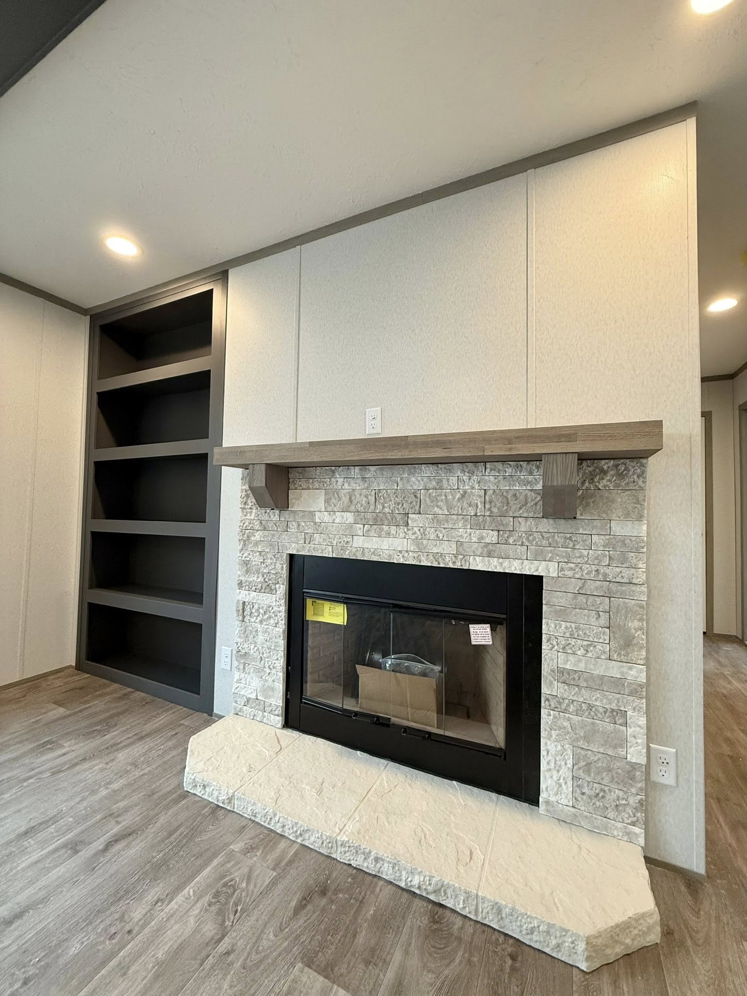 Modern living room with stone fireplace, wooden mantel, adjacent dark wood shelves, and light wood flooring. Soft lighting creates a cozy atmosphere.