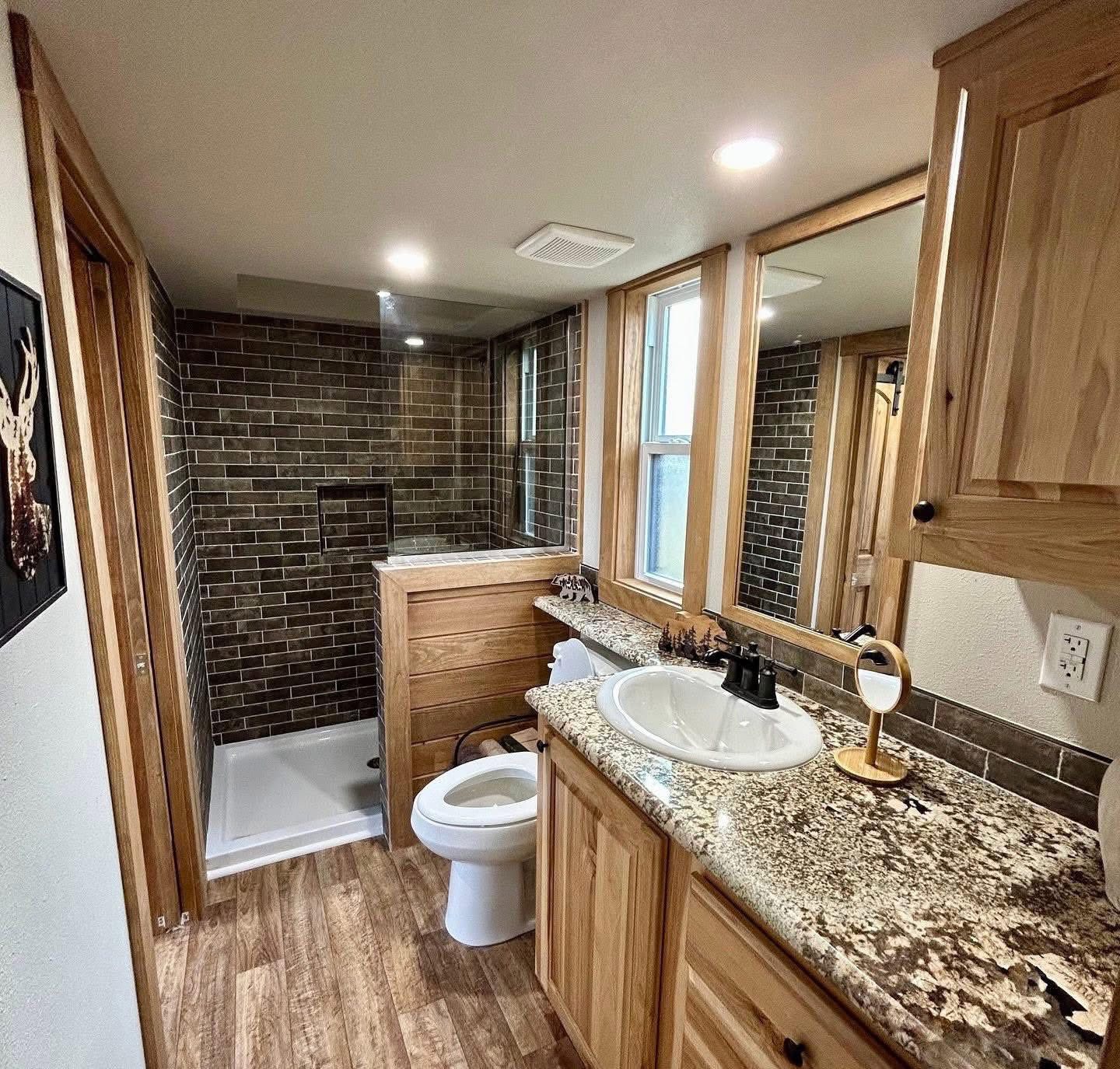Cozy bathroom with wood cabinetry, granite countertop, and a dark brick-tiled shower. Warm lighting and earth tones create a rustic ambiance.